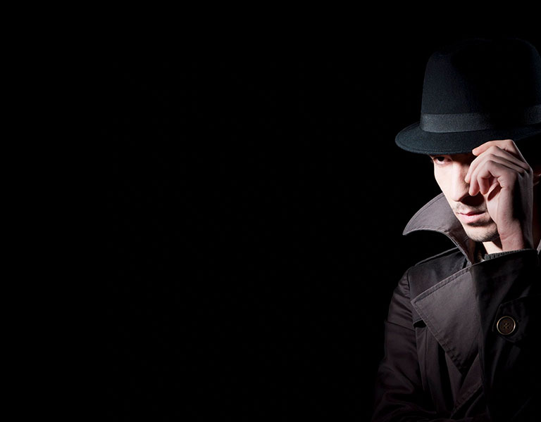 A man in a fedora and coat standing in the dark.